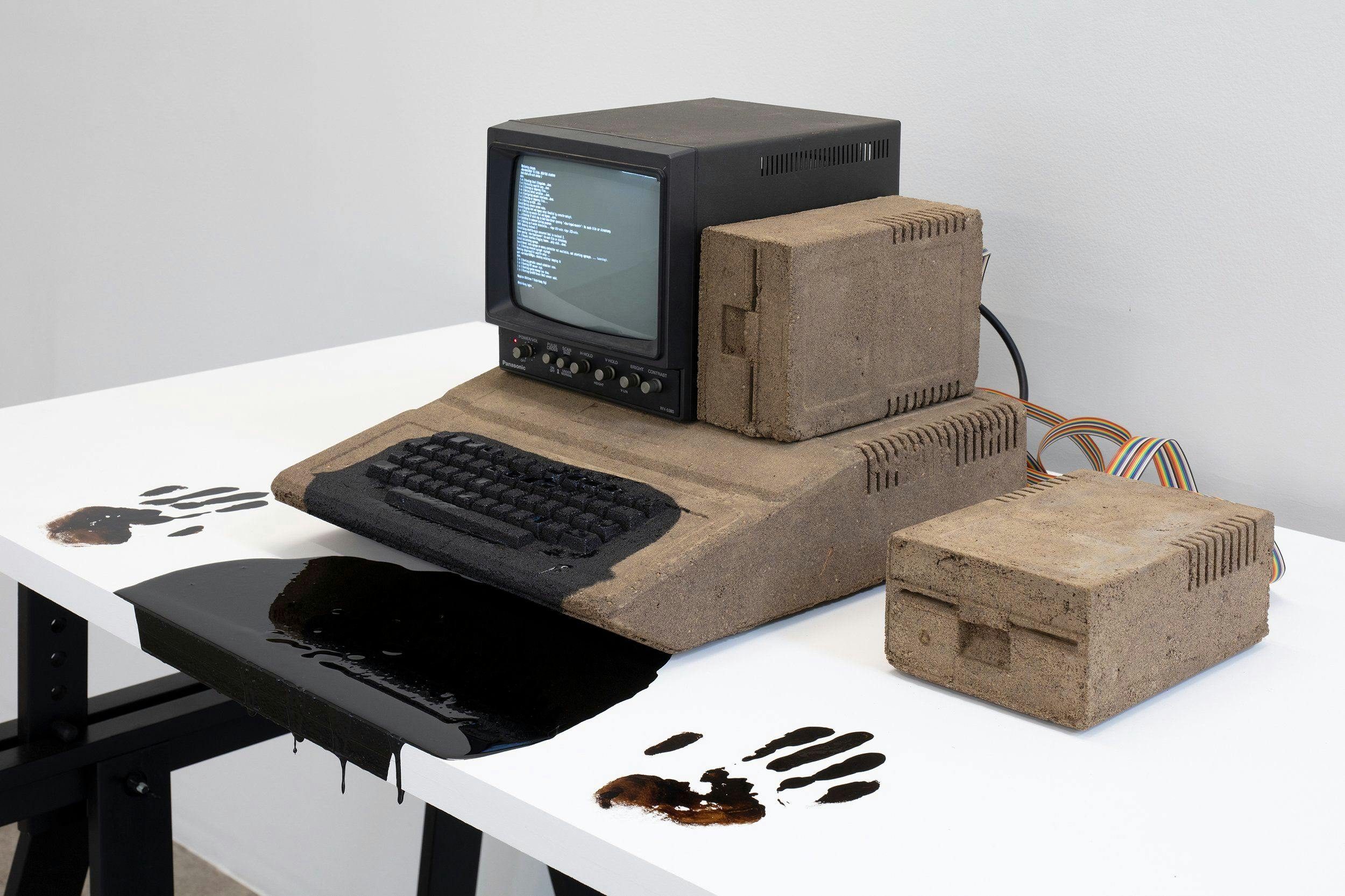 A small, vintage CRT monitor sits on top of a large, bulky keyboard and various computer parts cast in dirt, all atop of a sleek, white desk. There is a large spill of a gooey black liquid, on both the desk and the keyboard, as well as handprints with the same material on each side of the spill.