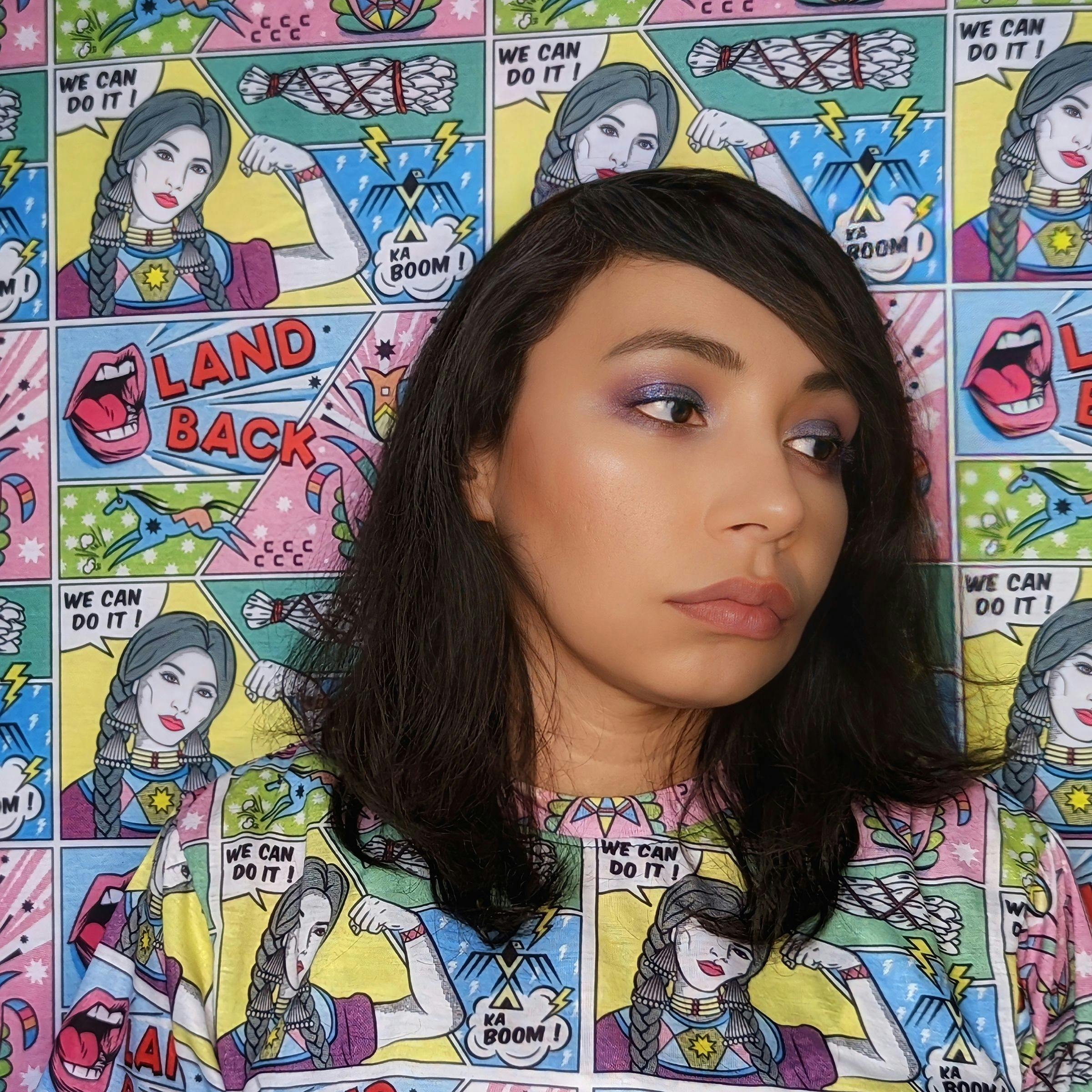 A Native American woman with brown skin, brown eyes, and shoulder-length brown hair looks off to the right at a 3/4th profile view. Her shirt and the background share the same colorful pattern, a comic book-style print of her own design, featuring a Native American woman flexing her arm with the phrases "We can do it" and "Land back."
