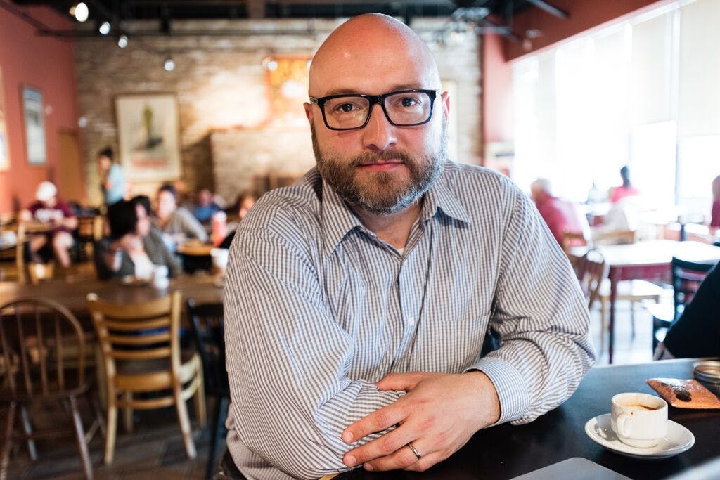 A fair-skinned man with a shaved head, cropped beard, and black glasses sits at a table in a cafe and looks into the camera. He wears a collared shirt with a gingham pattern.