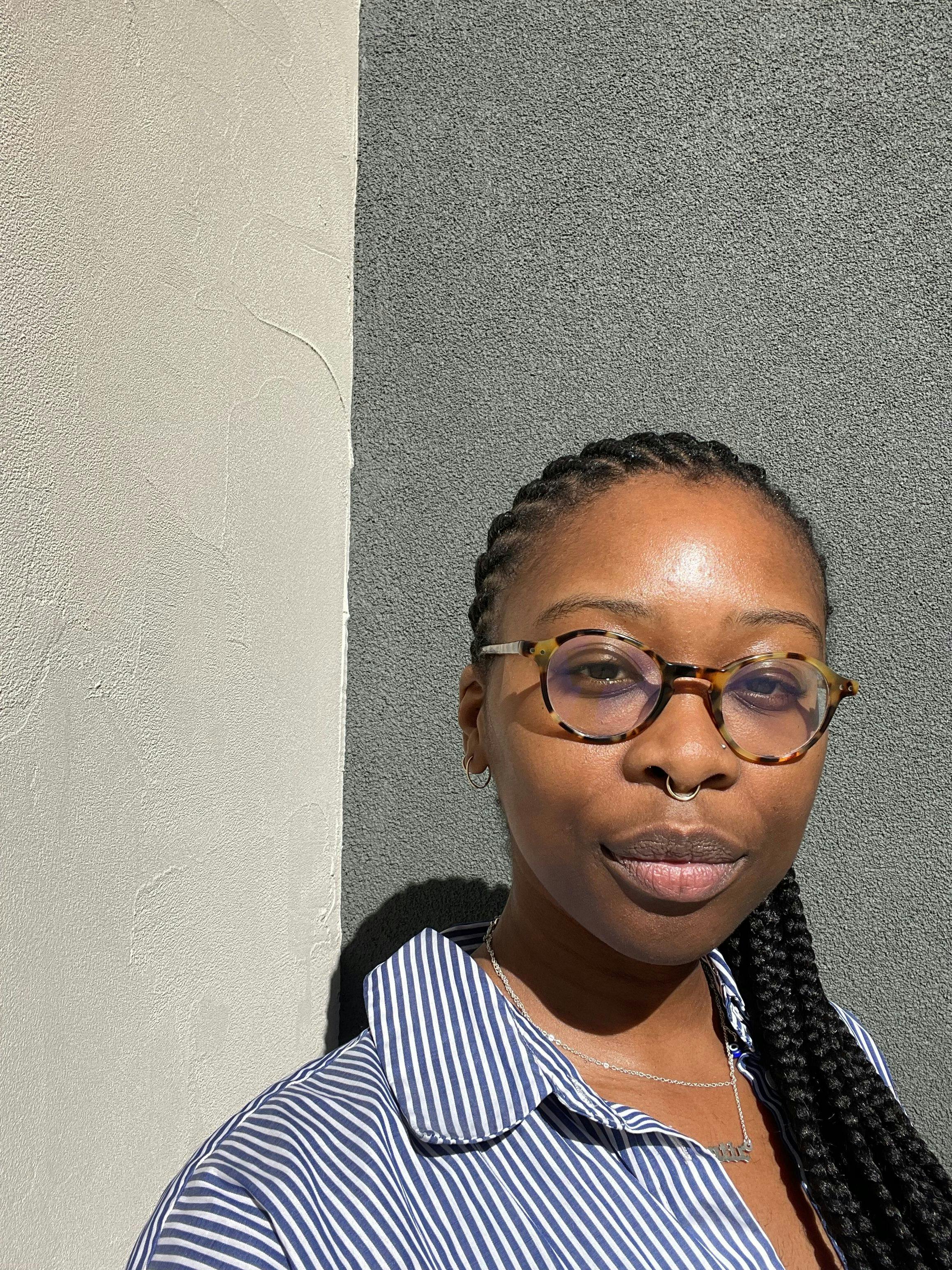 A black woman with tortoiseshell glasses, a septum piercing, and her hair in braids stands against a plaster wall under bright daylight and smiles softly to the camera.