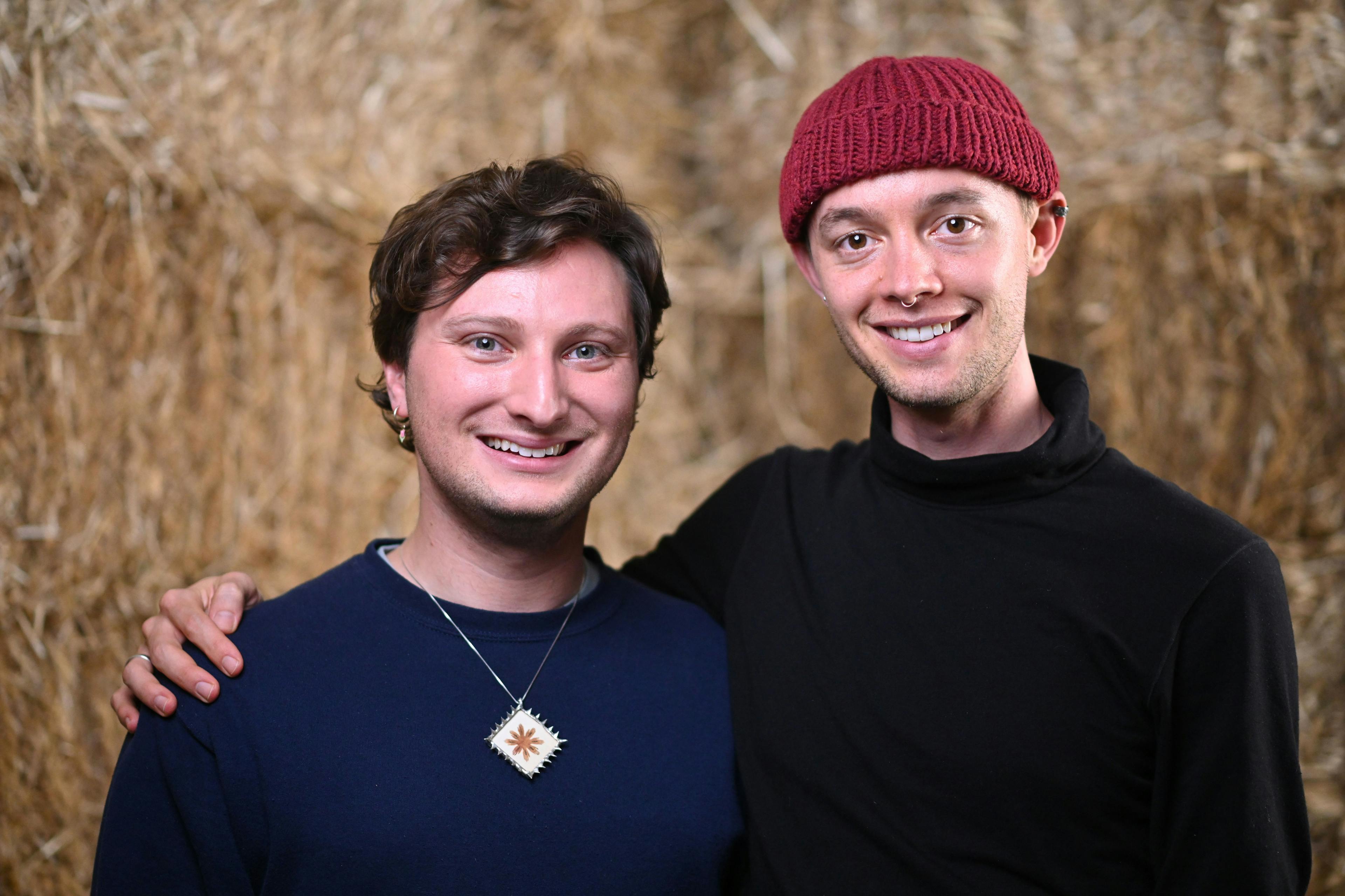 Two young white men smile in front of a blurry background resembling hay. One has wavy brown hair and wears a navy blue shirt and necklace, and the other wears a black turtleneck, red beanie, and a septum piercing.
