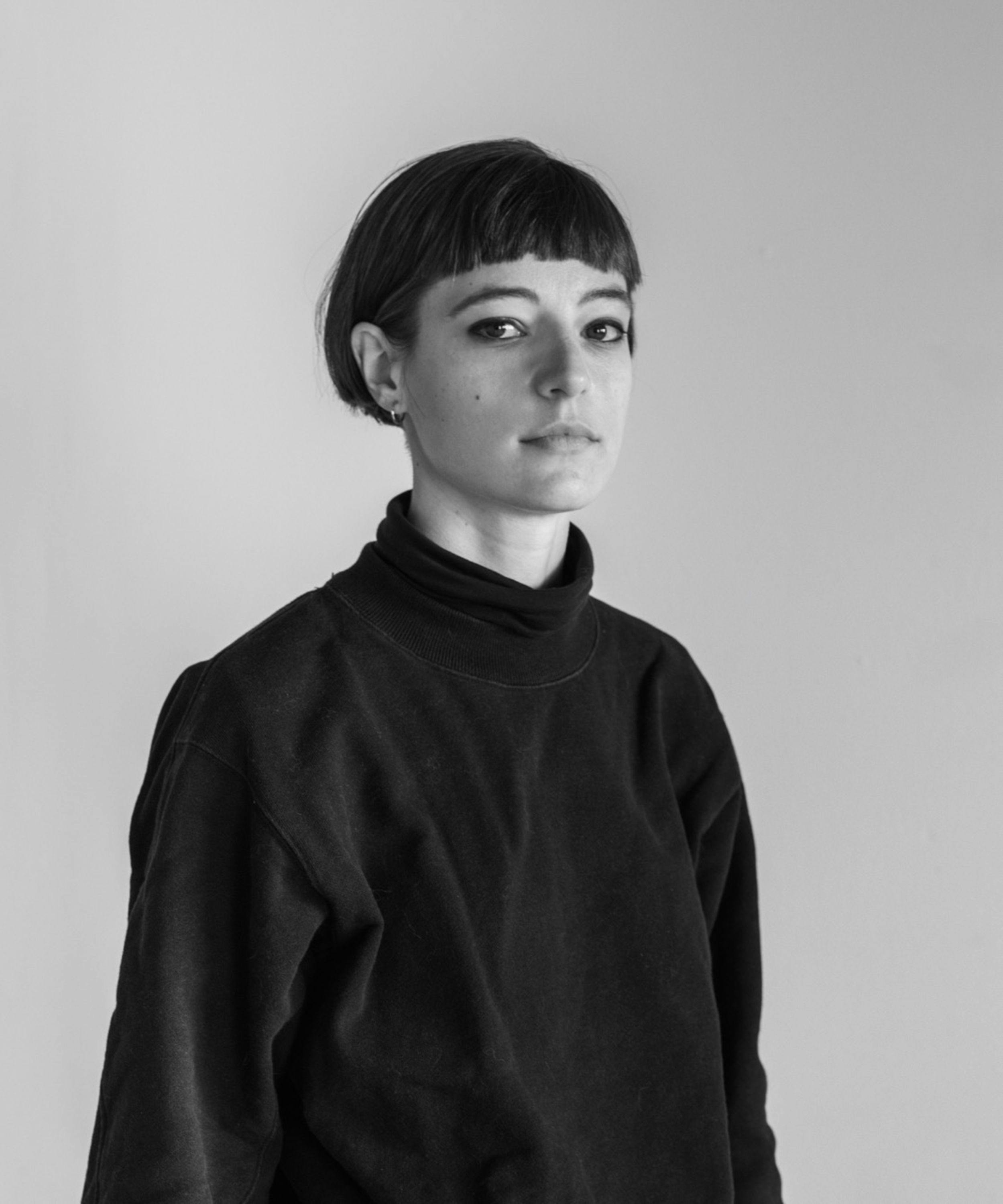 A black and white portrait of a white woman with a short bob haircut and bangs wearing a black turtleneck and small hoop earrings.