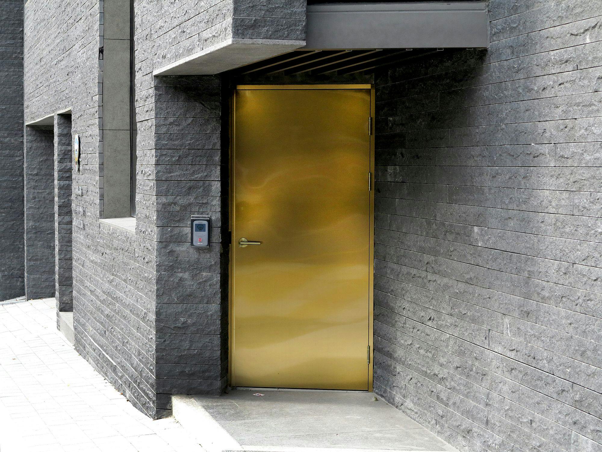A gold door with a minimal handle on the exterior of a gray building made of dark stone brick. To the left of the door is a mounted security keypad.