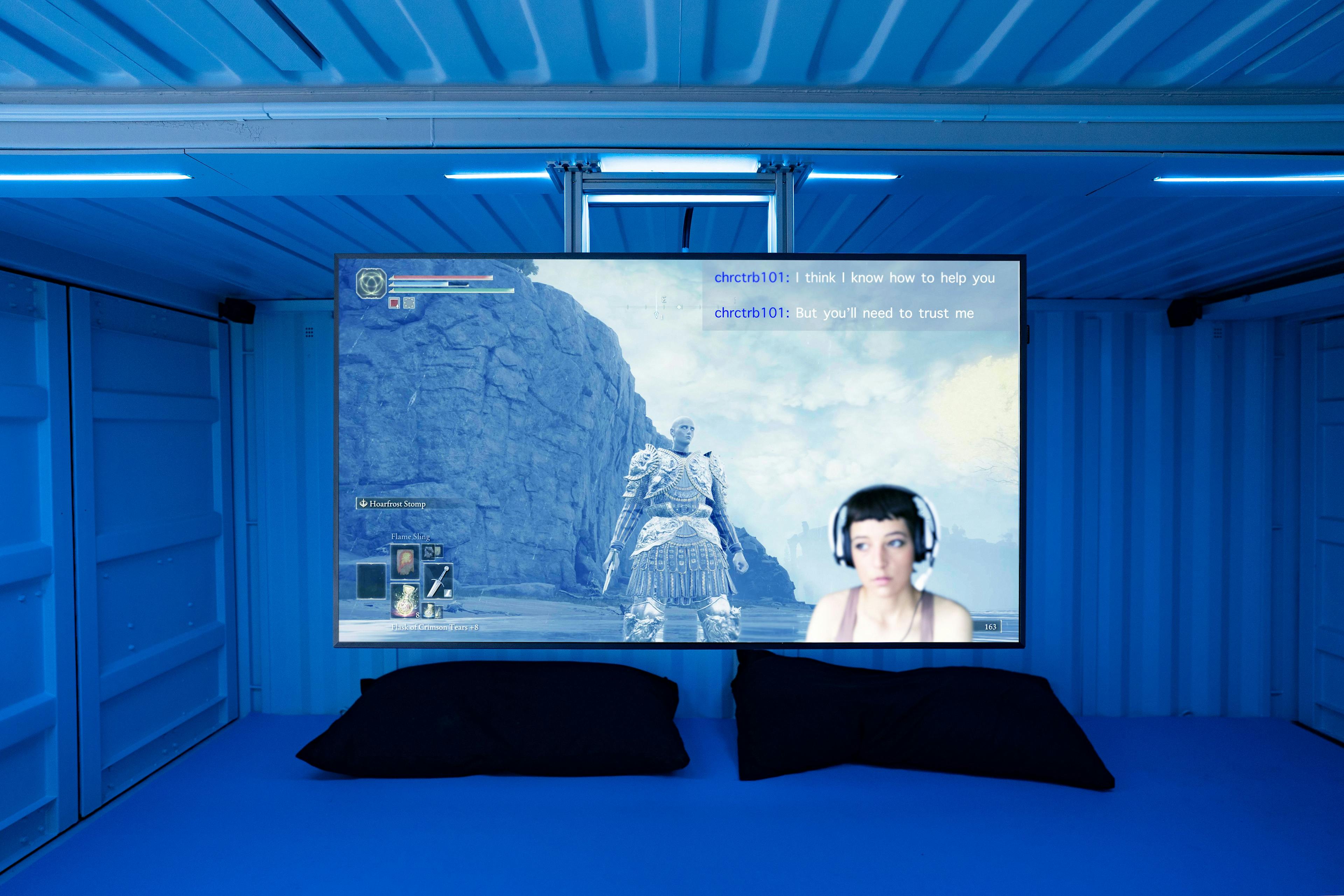 A ceiling-mounted monitor in a small blue room resembling a shipping container livestreams a video game. On the screen is an inset of a woman with a headset against a backdrop of a video game avatar in armor standing next to a large cliff. There are two dark pillows on the floor.
