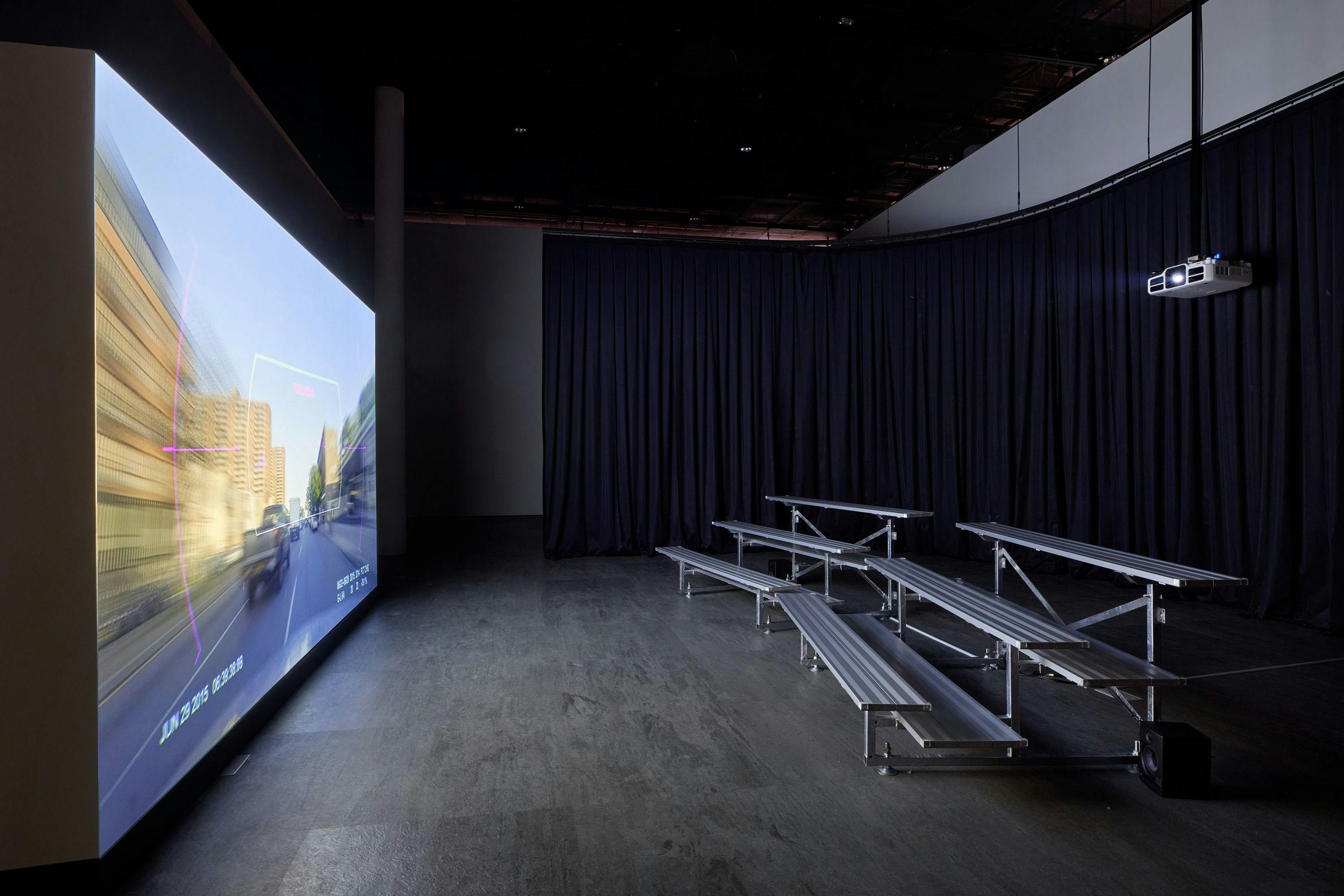 A dark, curtained room showcases a large projection on one wall and a set of bleachers facing it. The projection depicts dashcam footage, capturing cars driving ahead and buildings speeding by.