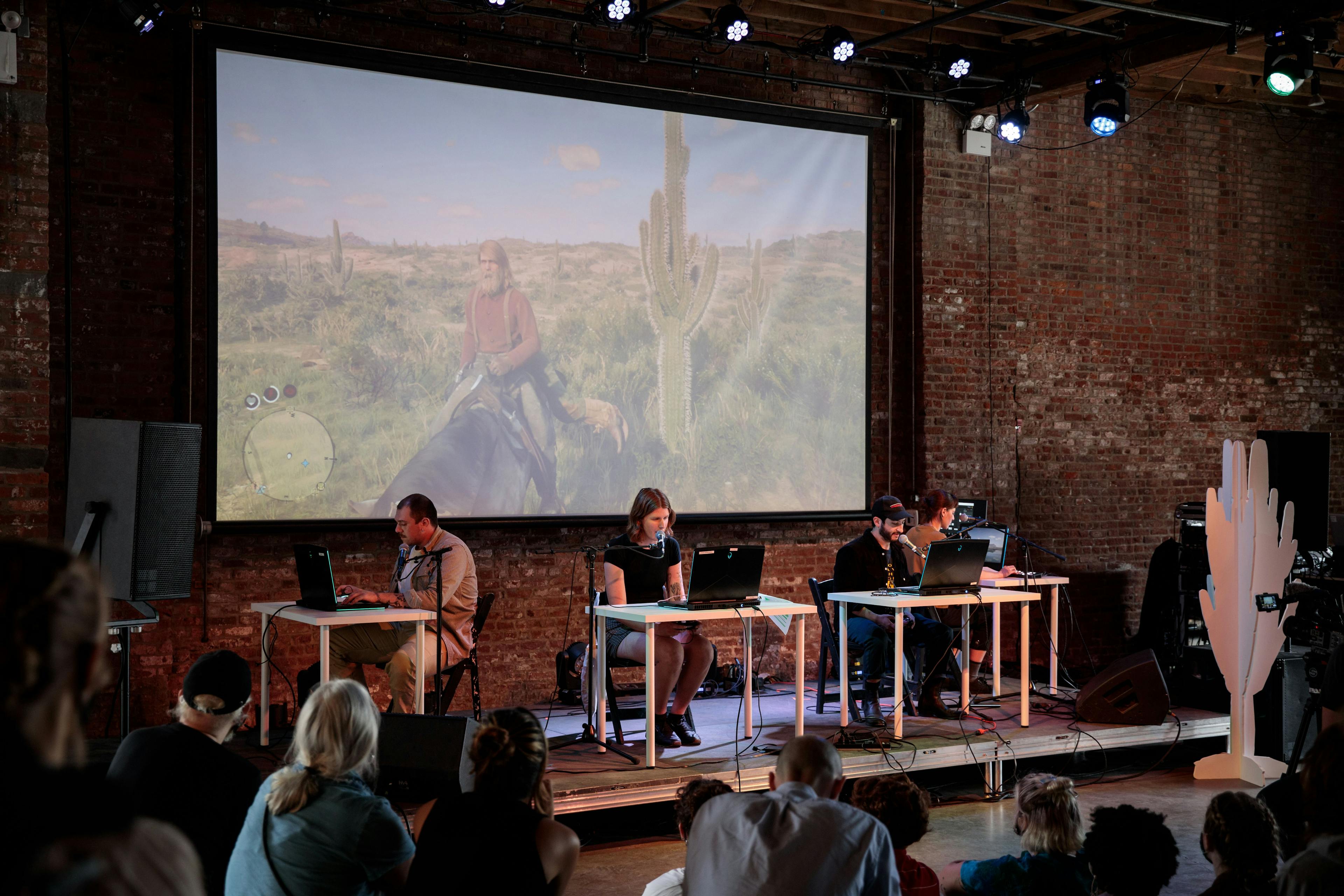 In a spacious brick room, four people sit at individual desks with laptops on a shallow stage, reciting out towards an audience on all three sides of the stage. Behind the performers is a large screen with a projection of a video game, showing a man on a horse in a desert scene.