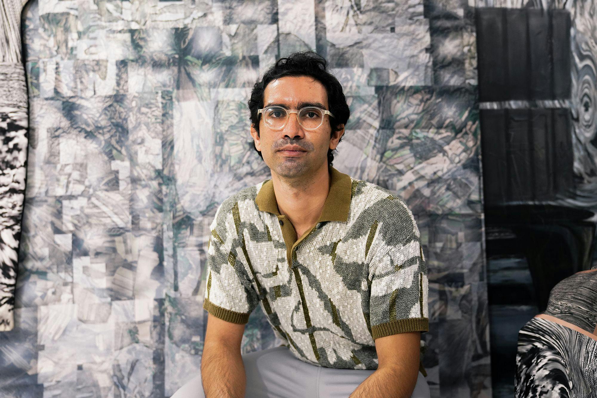 A portrait of a Latin American man with clear glasses and dark wavy hair, sitting in front of a patterned grayscale backdrop. He wears a woven shirt with an olive collar and a geometric design.