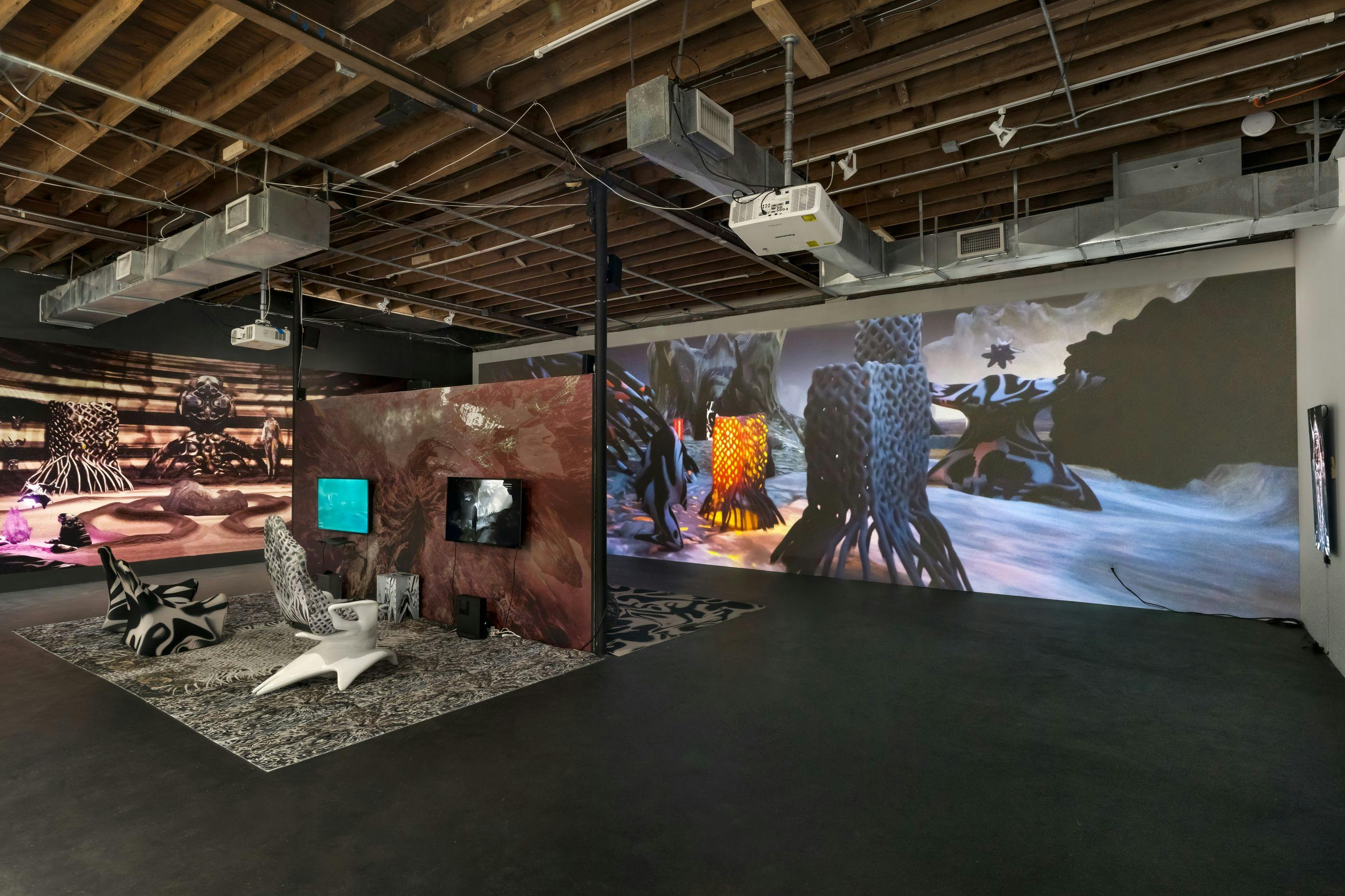 An installation view of a gallery show in a large room with tall ceilings: in the background is a wall-sized projection of a video game, depicting a surreal, abstracted landscape, and in the foreground is a temporary wall outfitted with an amorphous texture, television monitors, textured carpet, and angular seats.