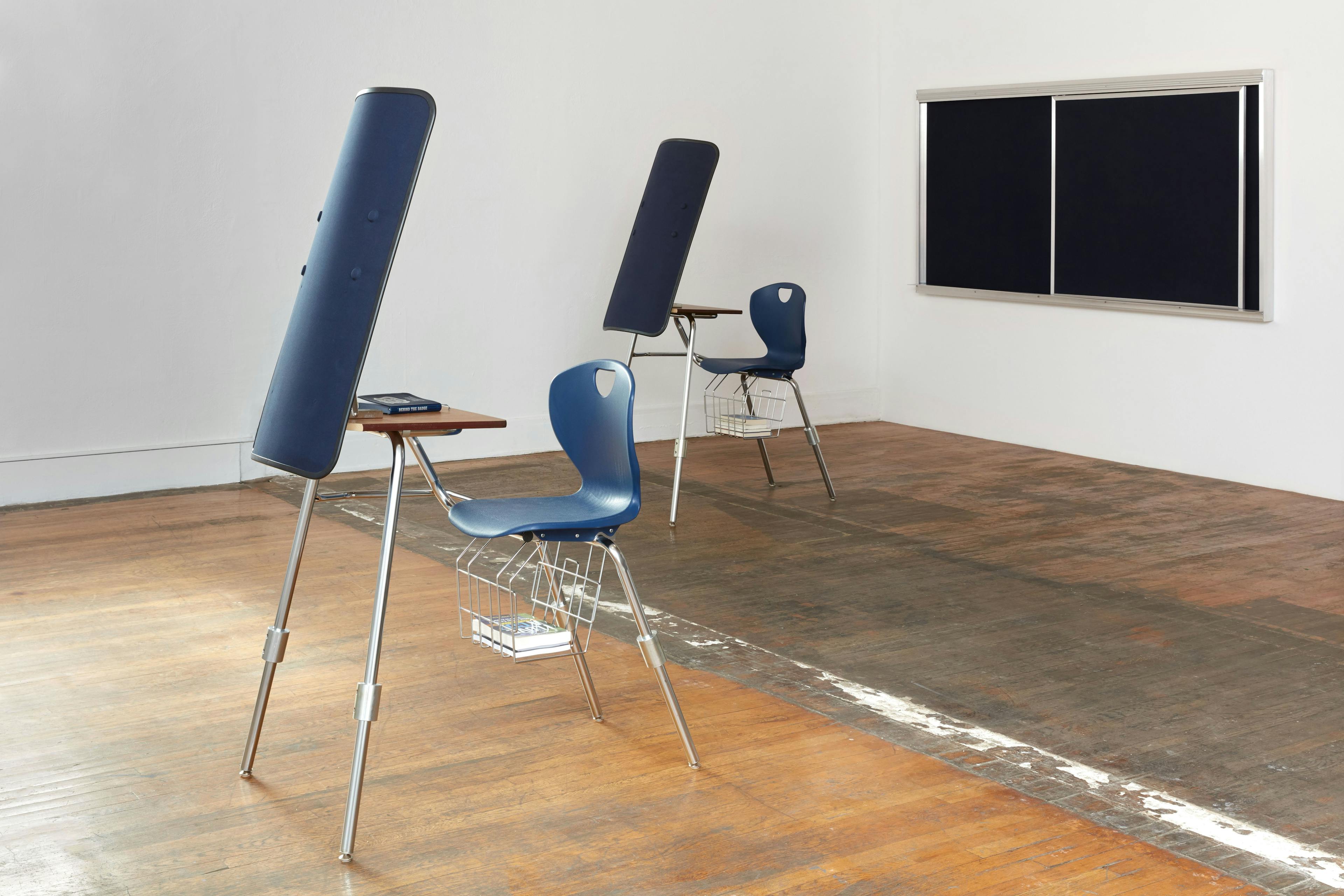 Two navy and wooden school desks, each modified to include a ballistic shield attached to the front, sit in a spacious white room with wooden floors. On the back wall is an empty chalkboard, and a small stack of books hangs below each desk.
