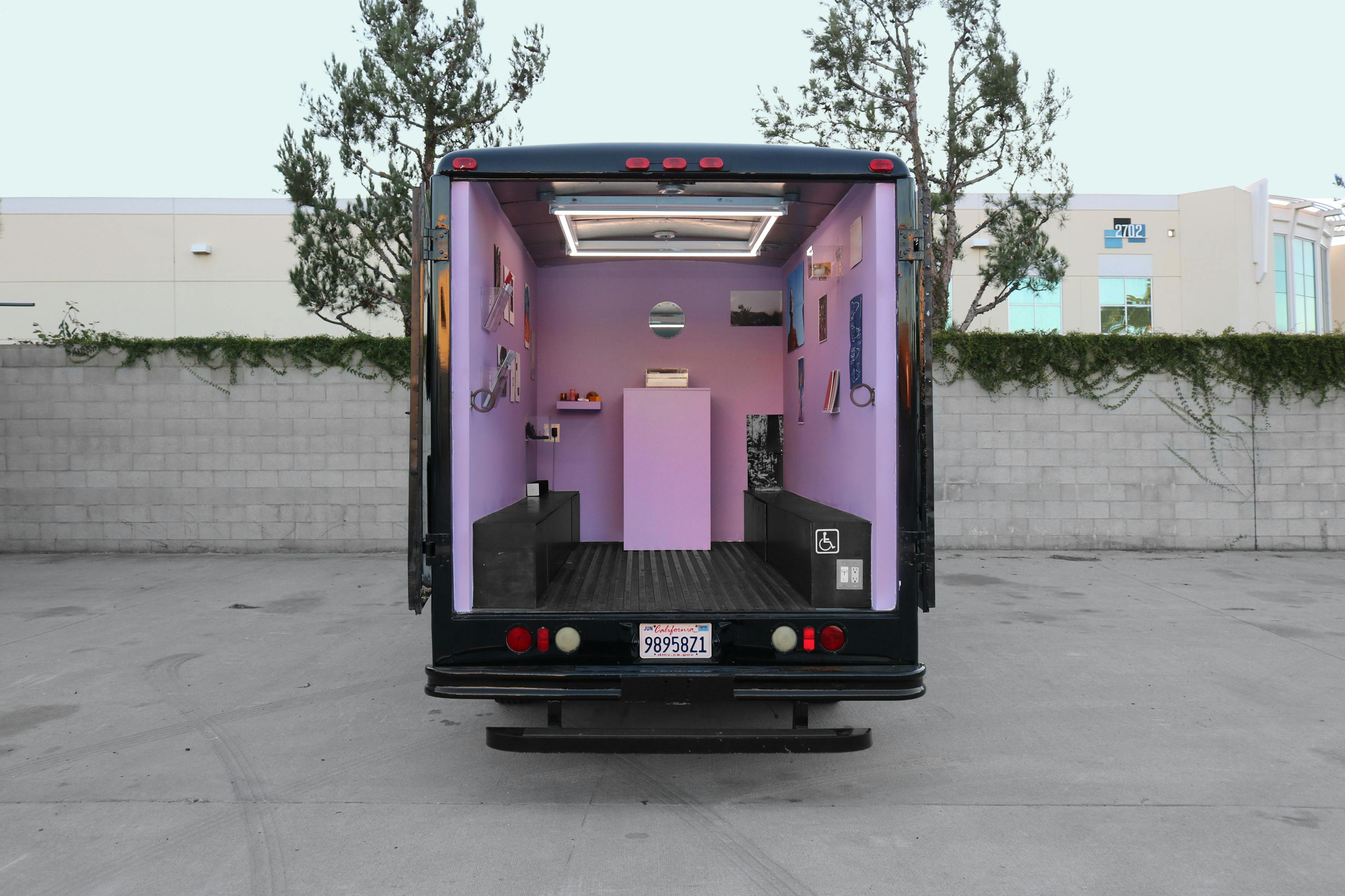 A small box truck sits parked in an outdoor lot, next to a cinderblock wall and two small trees. The truck's door has been rolled up, revealing a bright lavender interior filled with various small artworks on the walls and on a pedestal inside.