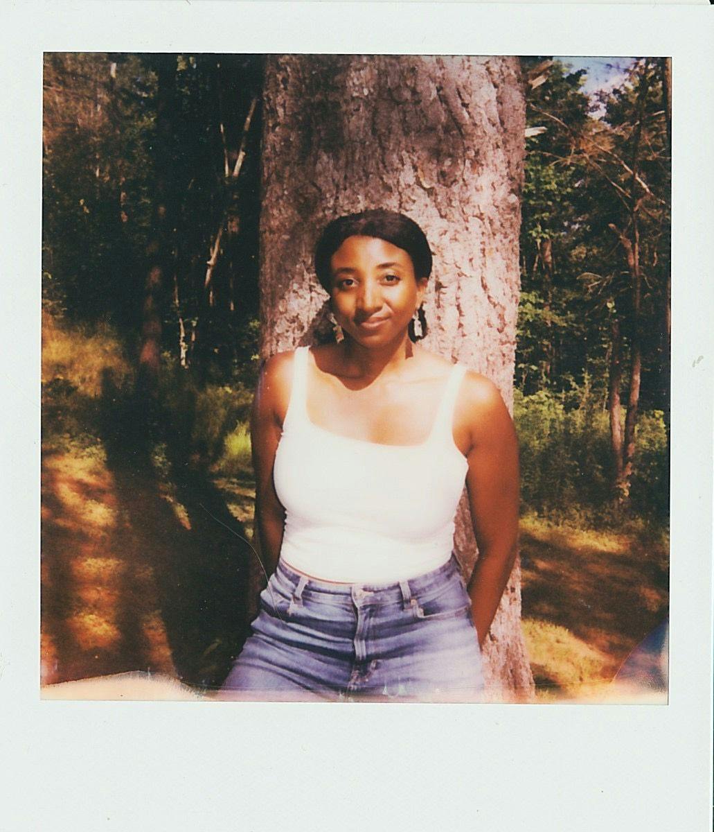 A polaroid photograph of a black woman with a center-part and low ponytail wearing a white tank top and jeans and leaning against a sun-dappled tree. The image is slightly blurry around the edges.