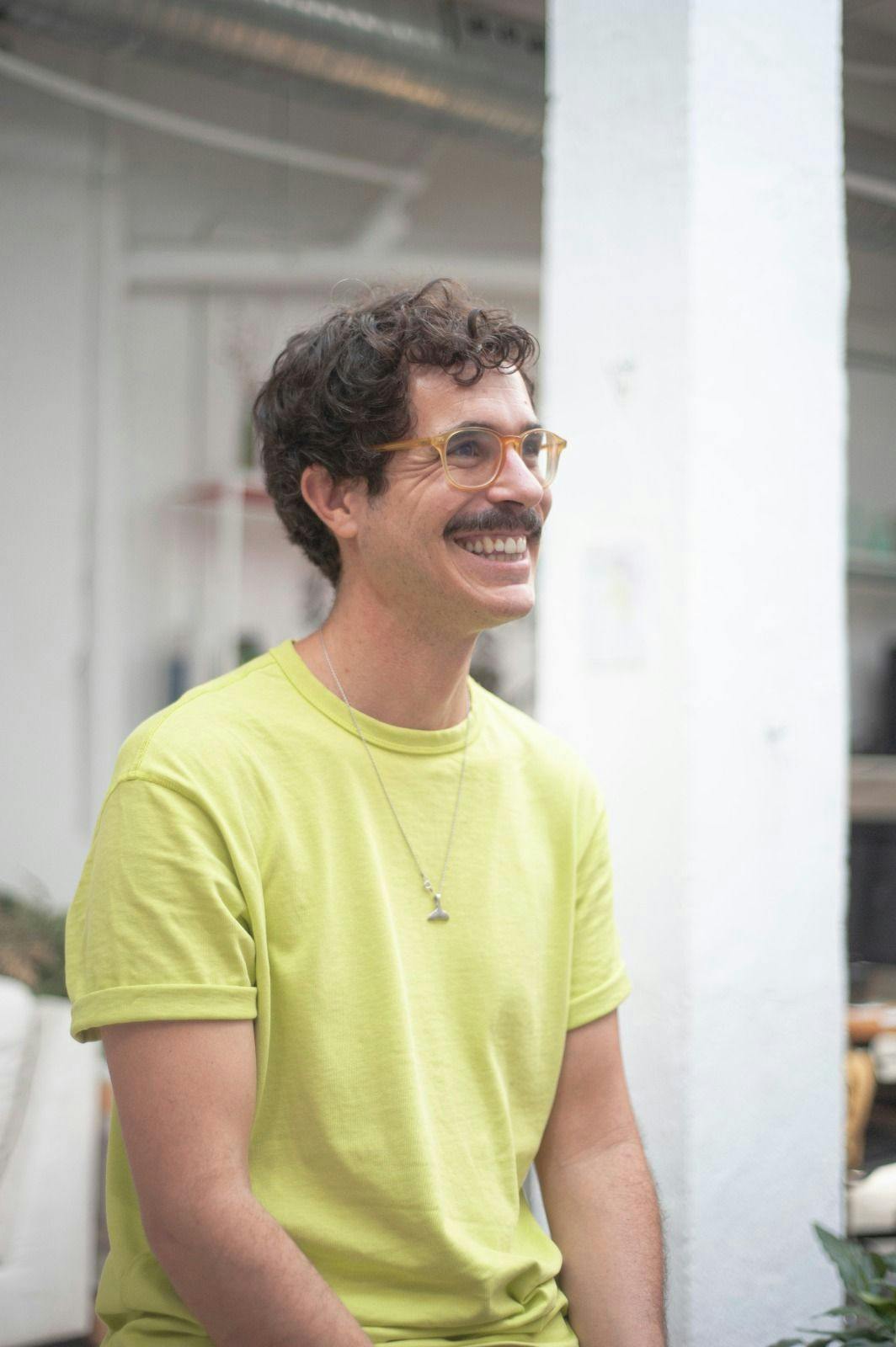 A man with light skin, curly brown hair, clear amber glasses, a mustache, and a pale lime green shirt sits in a bright, naturally lit room and looks off-camera, smiling.
