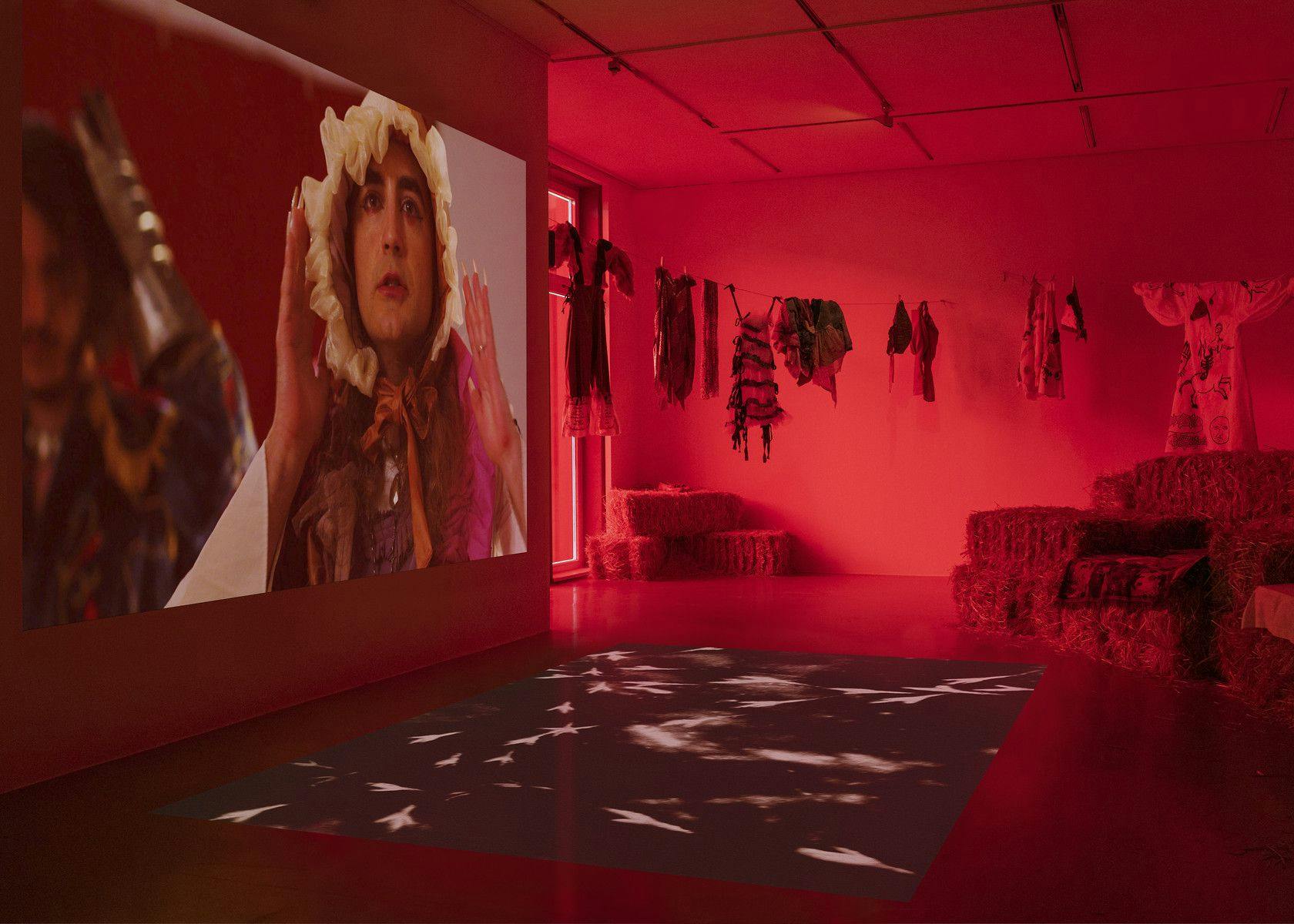 A dim, red-lit gallery space houses a large video projection on the wall of a person in a bonnet, a second projection on the floor of abstracted bird tracks, a clothesline of ruffly garments, and two large piles of hay bales.