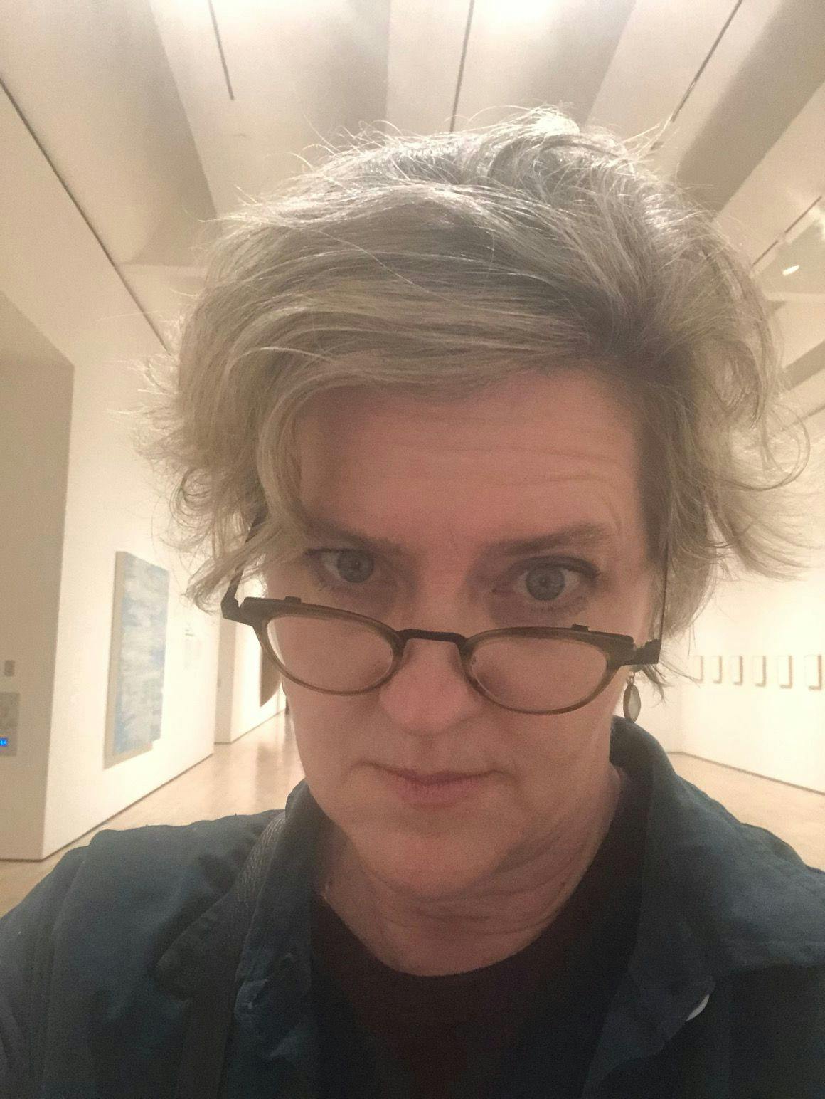 A white woman with silver hair looks over her wireframe glasses and into the camera. She wears a dark collared shirt, and stands within a warmly lit gallery space.